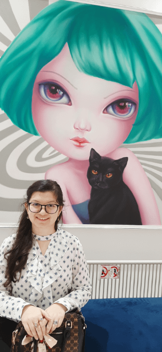 ragazza in posa davanti ad un quadro surrealista pop con una ragazza dal caschetto verde acqua con in braccio un gatto nero