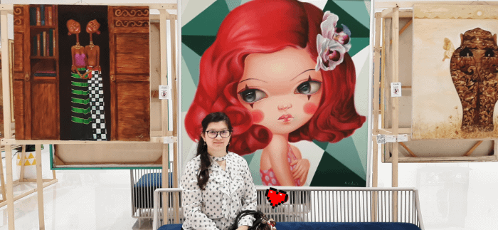 ragazza di fronte a un quadro surrealista pop con una ragazza dai capelli rossi e grandi occhi truccati con due linee verticali nere