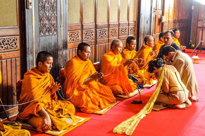 Monaci thailandesi seduti per terra su dei tappetini per essere ad un livello differente rispetto ai due sposi inchinati per terra di fronte a loro