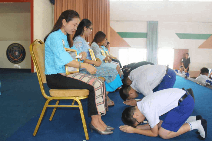 ragazzi thailandesi in divisa scolastica inginocchiati con il viso a terra davanti ai piedi delle proprie madri sedute su delle sedie
