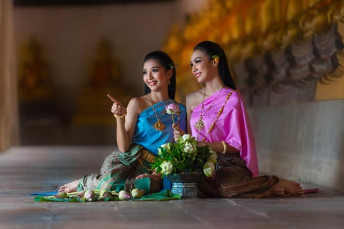 due ragazze thailandesi in costume tipico sedute per terra in un tempio, con le statue di Buddha disposte lungo il corridoio