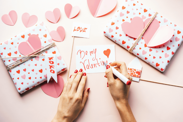 mani che scrivono un biglietto di san valentino in rosa, accanto a dei pacchetti rosa con cuori