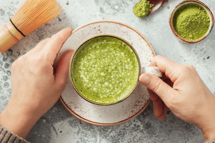 tazza di tè matcha vista dall'alto, con due mani che afferrano la tazza. sullo sfondo, in alto a sinistra lo strumento per fare il matcha, e in alto a destra una ciotola in legno con polvere di matcha
