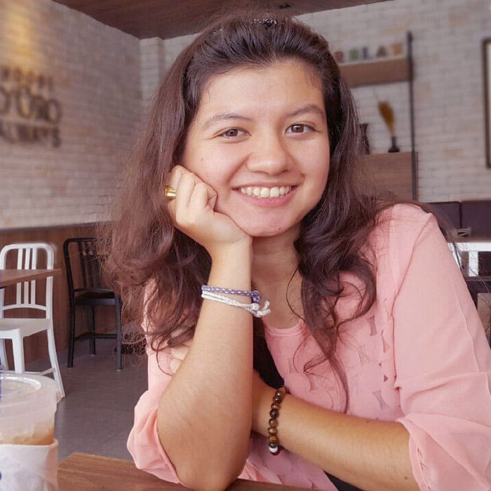 Ragazza sorridente con la guancia sinistra appoggiata sul palmo della mano, seduta ad un tavolino da bar