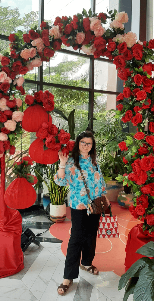 ragazza con occhiali, capelli semilunghi, con una vivace camicia verde acqua con rose arancioni, in piedi in posa sotto un arco decorato con rose rosse e rosa